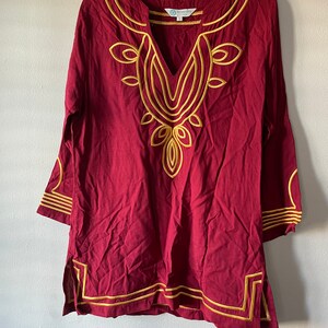 May include: A maroon tunic with gold embroidery around the neckline and hem. The tunic has a V-neck and three-quarter sleeves.