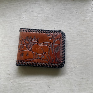 May include: A brown leather wallet featuring an embossed elk design set in a natural landscape. The wallet has a black braided border. The leather has a warm tone, and the design is intricately carved.