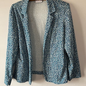 Op de afbeelding: Een tealblauwe blazer met een wit bloemenpatroon. De blazer heeft een kraag en lange mouwen. Het label op de blazer luidt "Flutterbye".
