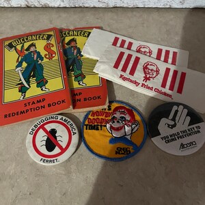 May include: A collection of vintage items including two red and yellow books with the word "Buccaneer" on the cover, a yellow and blue patch with the text "Howdy Doodie Timet", a red and white patch with the text "Kentucky Fried Chicken", a black and white button with a bug and the text "Debugging America Ferret", and a black button with a hand and the text "You hold the key to crime prevention Alberta Solicitor General".