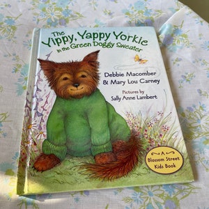 Puede incluir: Un libro infantil titulado "The Vippy, Yappy Yorkie in the Green Doggy Sweater" de Debbie Macomber & Mary Lou Carney. El libro presenta una ilustración de dibujos animados de un Yorkshire Terrier con un suéter verde.
