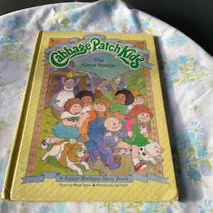 May include: A yellow book cover with the title "Cabbage Patch Kids The Great Rescue" and a picture of a group of Cabbage Patch Kids dolls and their animal friends. The book is a Parker Brothers Story Book, written by Mark Taylor and illustrated by Jan Brett.