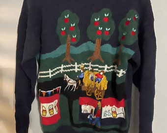 May include: Navy blue sweater with an embroidered pastoral scene. The design features apple trees with red apples, a horse-drawn cart, and figures near buildings. The sweater has a crew neck and long sleeves.