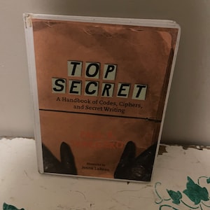 Puede incluir: Un libro titulado "Top Secret: A Handbook of Codes, Ciphers, and Secret Writing" con una cubierta marrón e ilustrado por Jenna LaReau.
