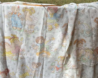 Op de afbeelding: Een wit stoffen laken met een herhalend patroon van Cabbage Patch Kids poppen in diverse outfits, waaronder tuinbroeken, jurken en jasjes, afgewisseld met kleine illustraties van bloemen, vlinders, bijen en regenbogen in zachte pastelkleuren.
