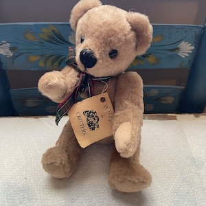 Puede incluir: Un oso de peluche marrón claro con ojos negros y nariz negra. El oso lleva una cinta de cuadros verdes y rojos alrededor del cuello. El oso está sentado sobre una superficie blanca y tiene una etiqueta que dice "Dolls & Critters".