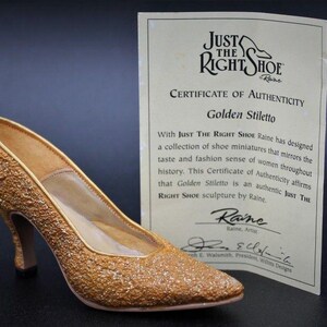 May include: A gold-coloured stiletto shoe sculpture with a textured finish. The shoe is positioned beside a certificate of authenticity from "Just The Right Shoe" by Raine. The certificate is labelled "Golden Stiletto".