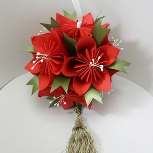 May include: A decorative, spherical ornament made from red paper flowers, green leaves, and white accents. It has a gold tassel and a white ribbon for hanging. This handcrafted item is ideal for festive decorations and adds a touch of elegance.