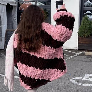 Può includere: Un cardigan a righe rosa e marrone lavorato a uncinetto con una texture soffice. Il cardigan è indossato da una persona con lunghi capelli castani.