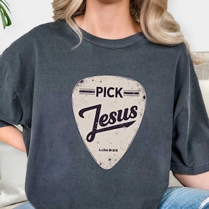 May include: Dark gray t-shirt featuring a guitar pick design with the words "PICK Jesus" in a vintage style. The text "Luke 9:23" is printed at the bottom of the pick. The shirt is a comfortable, casual wear item.