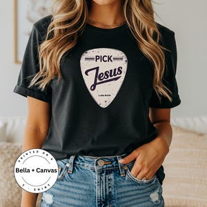 K&ouml;nnte beinhalten: Schwarzes T-Shirt mit einem Gitarrenplektrum-Design mit der Aufschrift "PICK Jesus" im Vintage-Stil. Das Shirt hat auch den Text "Lukas 9:23". Das Shirt ist auf Bella + Canvas gedruckt.