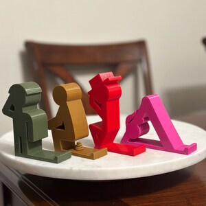 May include: Four phone stands in different colors and designs. The stands are made of plastic and are shaped like people in different poses. The stands are on a white marble surface.