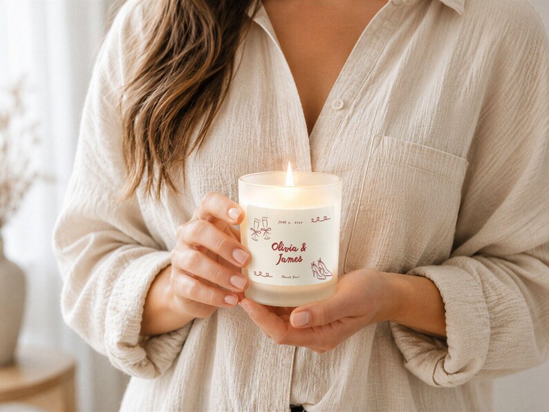 May include: A lit candle in a frosted glass jar. The jar has a label with the names "Olivia & James" and decorative illustrations. The candle is held by a person wearing a beige button-down shirt.