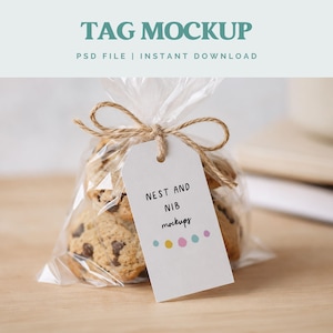Pode incluir: Um saco de celofane transparente, amarrado com barbante, contendo biscoitos com pepitas de chocolate. Uma etiqueta branca com o texto "NEST AND NIB mockups" e pontos coloridos pende do barbante. As palavras "TAG MOCKUP" e "PSD FILE | INSTANT DOWNLOAD" estão no topo.