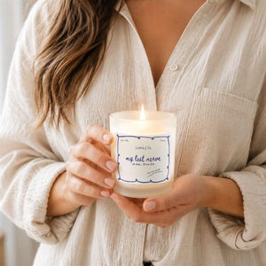 May include: A lit, frosted glass candle with a blue and white label that reads "my last nerve." The candle is held by a person wearing a beige button-down shirt. The candle is in focus, with a soft, warm glow.