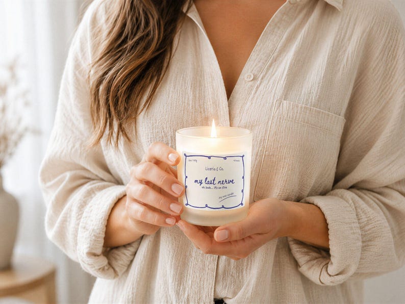May include: A lit, frosted glass candle with a blue and white label that reads "my last nerve." The candle is held by a person wearing a beige button-down shirt. The candle is in focus, with a soft, warm glow.