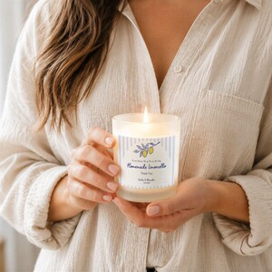 May include: A lit, frosted glass candle with a label that reads "Homemade Limoncello" and features a blue and white striped design with lemon illustrations. The candle is held in front of a beige shirt.