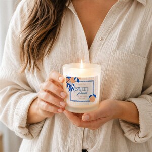 May include: A lit, frosted glass candle with a label that reads "Sweet Peach" is held in front of a person. The label has a blue border with peach and leaf illustrations. The candle is in a clear glass container.