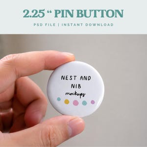 Może przedstawiać: Biała przypinka o średnicy około 5,7 cm, trzymana między palcami. Na guziku znajduje się czarny napis "NEST AND NIB mockups" z rzędem kolorowych kropek poniżej. Na górze obrazu widnieje napis "2.25" PIN BUTTON".