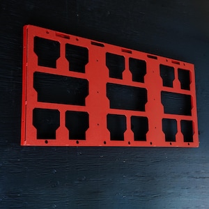 May include: A rectangular, bright orange metal organiser panel featuring a grid of various sized cutouts. The flat, rigid frame has a textured finish and includes multiple small circular holes along the edges for mounting or attachment purposes.