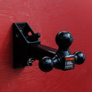 Peut inclure: Un support de boule d'attelage Curt noir doté d'une conception à trois boules de tailles distinctes. Le support est fixé à une bride de montage noire. Une étiquette sur l'attelage affiche les capacités de poids en kilogrammes pour chaque boule.