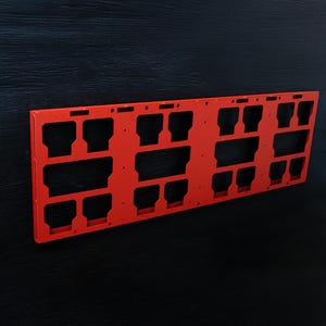 May include: A rectangular red plastic frame featuring a grid of various sized cutouts and slots. The flat, structured panel has a series of symmetrical openings arranged in a repeating pattern across its surface, designed for mounting components.