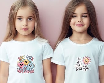 Op de afbeelding: Twee meisjes dragen witte T-shirts met verschillende ontwerpen. Het meisje links draagt een T-shirt met de tekst "Self Care is Not Selfish" en een cartoonillustratie van een hart, een smileygezicht en een vierkant. Het meisje rechts draagt een T-shirt met de tekst "The Sun Will Shine Again" en een cartoonillustratie van een zon en een wolk.