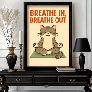 May include: Framed art print featuring a cartoon cat meditating on a green mat. The cat is brown and tan with its eyes closed. The words "BREATHE IN, BREATHE OUT" are printed in orange above the cat. The print is in a black frame.