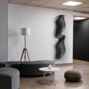 May include: A modern interior scene featuring a black, abstract wall sculpture with a layered design. A wooden tripod floor lamp with a white lampshade stands near a gray sectional sofa and a small white table.