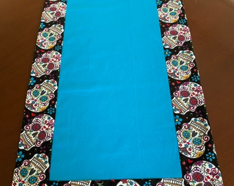 May include: A turquoise table runner with a black and white border featuring a repeating pattern of colorful sugar skulls.