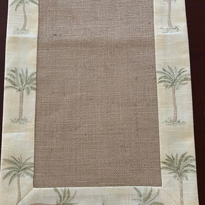 May include: A rectangular table runner with a natural burlap center and a patterned border. The border features a repeating design of palm trees in shades of green and brown against a cream-colored background. The runner is designed for home decor.