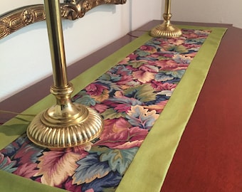 May include: A green table runner with a floral pattern and a gold trim. The runner is on a wooden table with two gold candle holders.