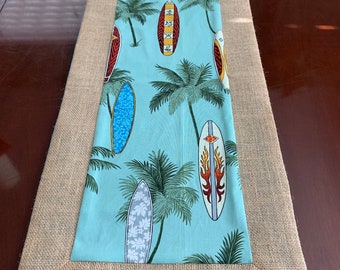 May include: A rectangular table runner with a light blue center featuring surfboards and palm trees. The center is framed by a burlap border. The surfboards are in various colors and designs.