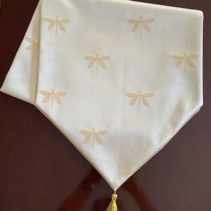 Puede incluir: Un camino de mesa blanco con un diseño de libélula bordado en oro y una borla dorada.