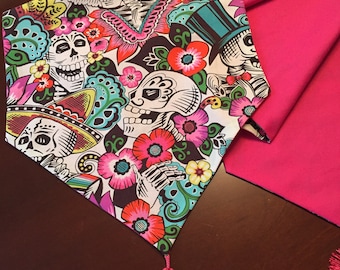 May include: A pink and white table runner with a Day of the Dead theme. The runner features colorful skulls, flowers, and butterflies. It has a pink tassel on each end.