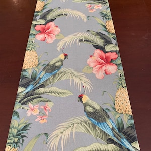Peut inclure: Un chemin de table gris avec un imprimé tropical représentant des perroquets, des ananas et des fleurs d'hibiscus roses.