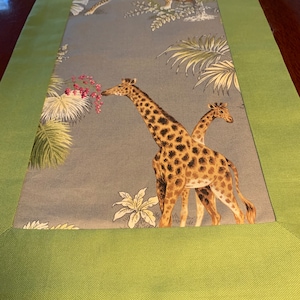 May include: A green table runner with a gray center panel featuring a printed design of giraffes and a leopard in a jungle setting. The design includes palm trees, leaves, and flowers.