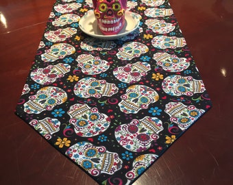 May include: A black table runner with a repeating pattern of colorful sugar skulls and flowers. A small succulent plant is in a red sugar skull planter on top of the runner.
