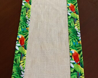 May include: A beige table runner with a green, yellow, and blue border featuring tropical leaves and parrots.