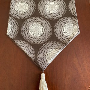 May include: A brown table runner with a white geometric pattern of circles. The runner has a white tassel at the end.