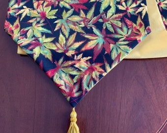 May include: A black table runner with a pattern of colorful autumn leaves. The runner has a gold tassel at the end and is made of a soft, luxurious fabric.