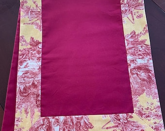 Può includere: Runner da tavolo rettangolare bordeaux con bordo giallo e motivo toile de Jouy rosso. Il runner in tessuto è piegato, mostrando i colori contrastanti e il design. Le dimensioni del runner non sono visibili.