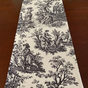 May include: A black and white toile table runner with a French country design featuring people and trees. The runner is made of fabric and has a cream background.