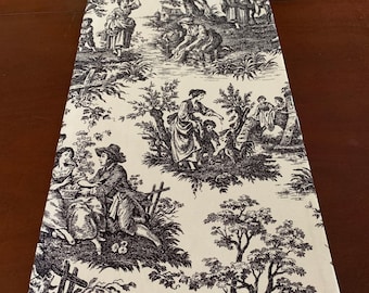 May include: A black and white toile table runner with a French country design featuring people and trees. The runner is made of fabric and has a cream background.
