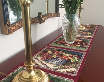 May include: A red table runner with a white lace border features a repeating pattern of fruit baskets. The runner is decorated with a variety of fruits, including apples, grapes, pears, and plums. A bouquet of yellow, orange, and red flowers in a clear glass vase sits on the runner.