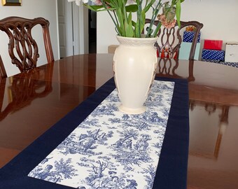 May include: A blue and white toile table runner with a navy blue border. The runner is on a wooden table with a vase of pink and white flowers.