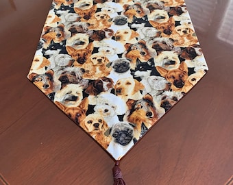 May include: A white table runner with a brown tassel. The runner is covered in a pattern of dog faces in various breeds and colors.