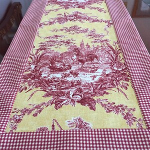 May include: A rectangular table runner with a yellow center featuring a red toile design of a rooster, buildings, and floral elements. The border is a red and white gingham pattern. This decorative runner is perfect for a dining table.