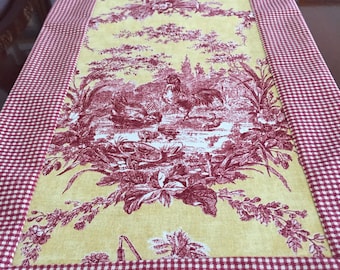 May include: A rectangular table runner with a yellow center featuring a red toile design of a rooster, buildings, and floral elements. The border is a red and white gingham pattern. This decorative runner is perfect for a dining table.