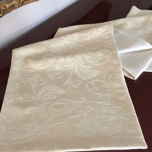 Op de afbeelding: Crèmekleurige tafelloper en servetten met een reliëf bloemmotief. De loper is opgevouwen en ligt op een donker houten oppervlak, met verschillende gevouwen servetten in de buurt. De stof heeft een subtiele glans.